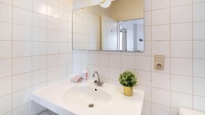 Een badkamer met witte betegelde wanden, een witte wastafel, een spiegel, een waterkraan, een kleine potplant en een handzeepfles.