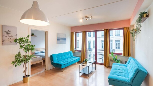 Een moderne woonkamer met een blauwe bank, een glazen salontafel en een groot raam met gordijnen.
