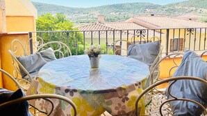 A round table with a floral tablecloth, surrounded by metal chairs with cushions, on a balcony with a view of green hills and red-roofed buildings. - New Orange Studio Apartment in Limpiddu, Budoni – Pool, Terrace, Wi-Fi (Budoni)