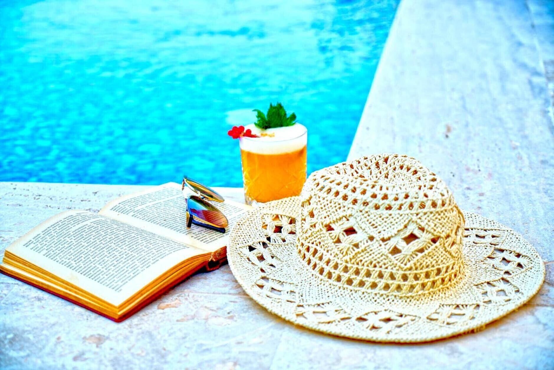 An open book, a straw hat, and a glass of drink by a poolside.