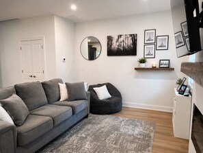 Ein modernes Wohnzimmer mit einem grauen Sofa, einem Sitzsack, einem runden Spiegel und gerahmten Bildern an der Wand.
