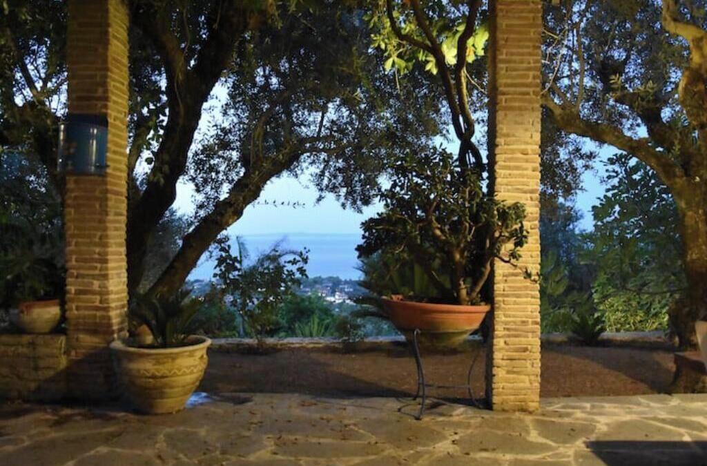 Un espacio al aire libre cubierto con pilares de ladrillo, plantas en macetas y vistas al mar.