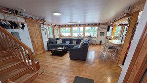 A spacious living area with a wooden floor, a sofa, a coffee table, a staircase, and a dining area with chairs and a table. - 3-Bedroom House on Beautiful Flying Pond in Mt. Vernon, ME (Mount Vernon)