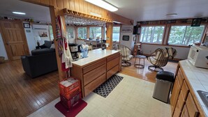 A cozy kitchen with wooden cabinets, a dining area with chairs, and a view through large windows. - 3-Bedroom House on Beautiful Flying Pond in Mt. Vernon, ME (Mount Vernon)