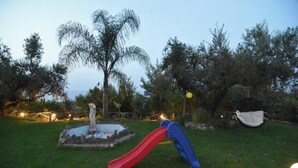 Un parque infantil con un tobogán rojo y una estructura de escalada azul, rodeado de árboles y un pequeño estanque.