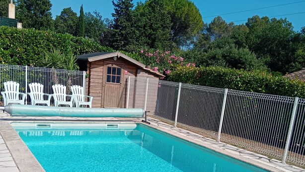 Una piscina con borde de piedra, una cabaña de madera, sillas blancas y una zona cercada.