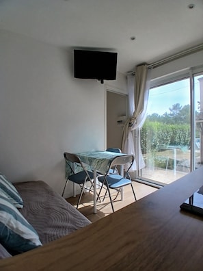 Una habitació amb un llit, una taula i cadires, una televisió penjada a la paret i una porta corredissa de vidre que dona a una zona exterior.