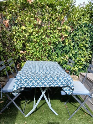 A table with a blue and white patterned tablecloth, surrounded by folding chairs, set outdoors with a hedge in the background. - Quiet apartment with pool (Grasse)