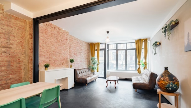 A modern living room with a brick wall, a sofa, a dining table with chairs, and a large vase on a side table. - Spacious 2BR Stay for 14 by Grand Central (Antwerp)