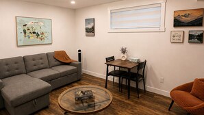 A living room with a sectional sofa, a glass coffee table, a dining table with chairs, and a window with blinds. - Private Suite w/ Full Kitchen Near Banff National Park (Cochrane)
