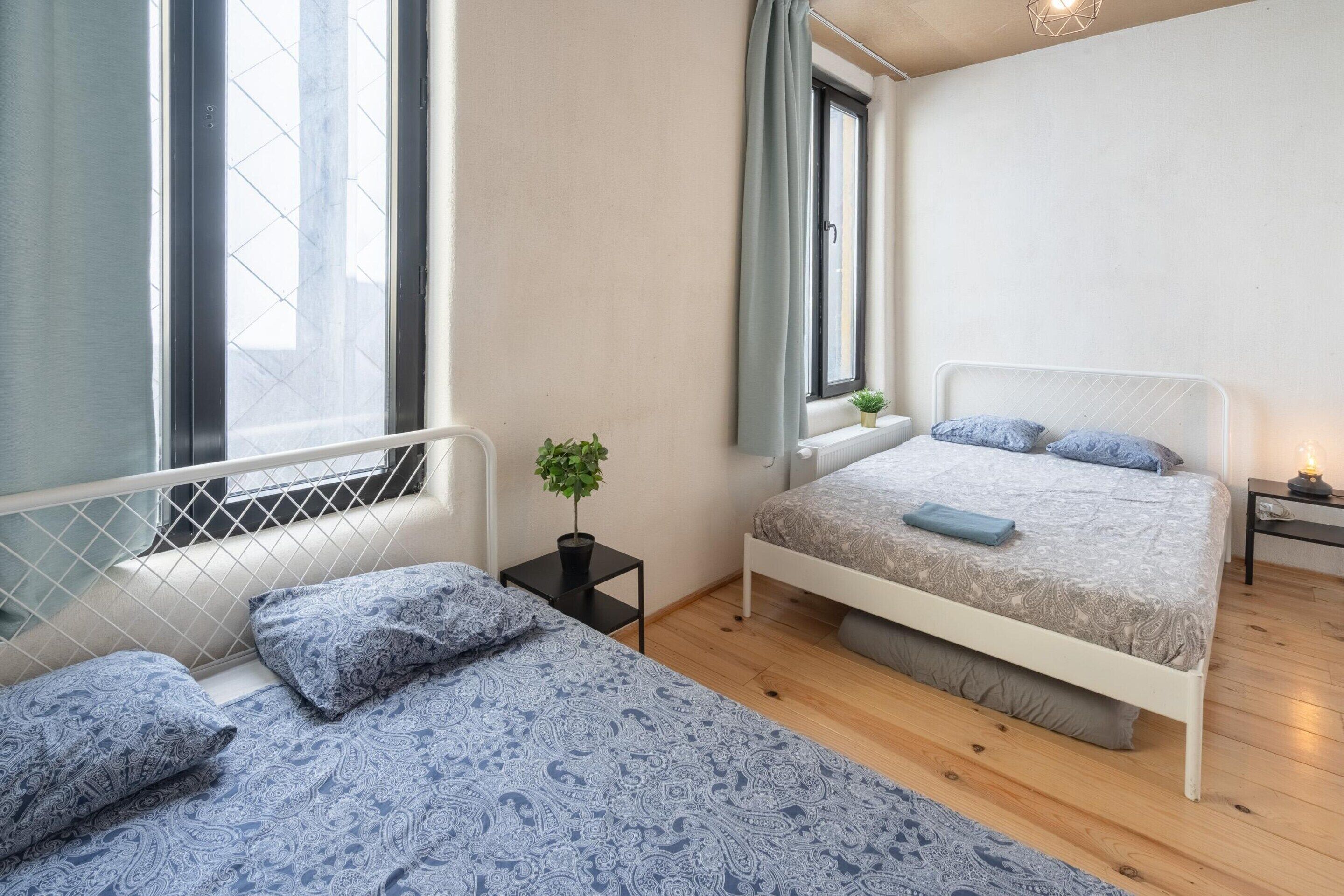 Une chambre avec deux lits, une plante en pot, une table de chevet et une fenêtre avec des rideaux.