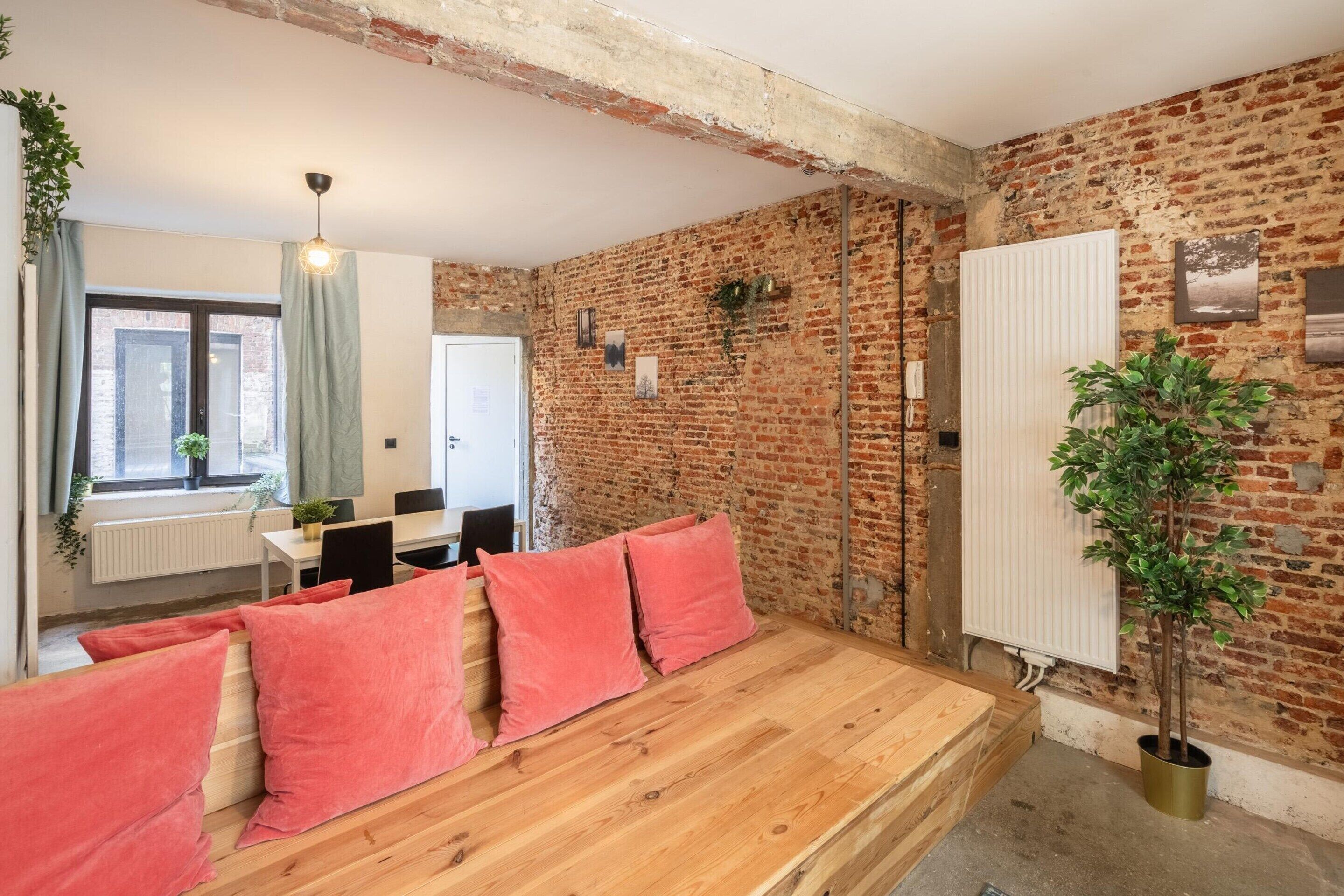 Una habitación con paredes de ladrillo visto, un banco de madera con cojines rojos, un radiador y una planta en maceta.