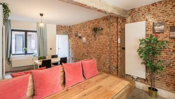 Una habitación con paredes de ladrillo visto, un banco de madera con cojines rojos, un radiador y una planta en maceta.