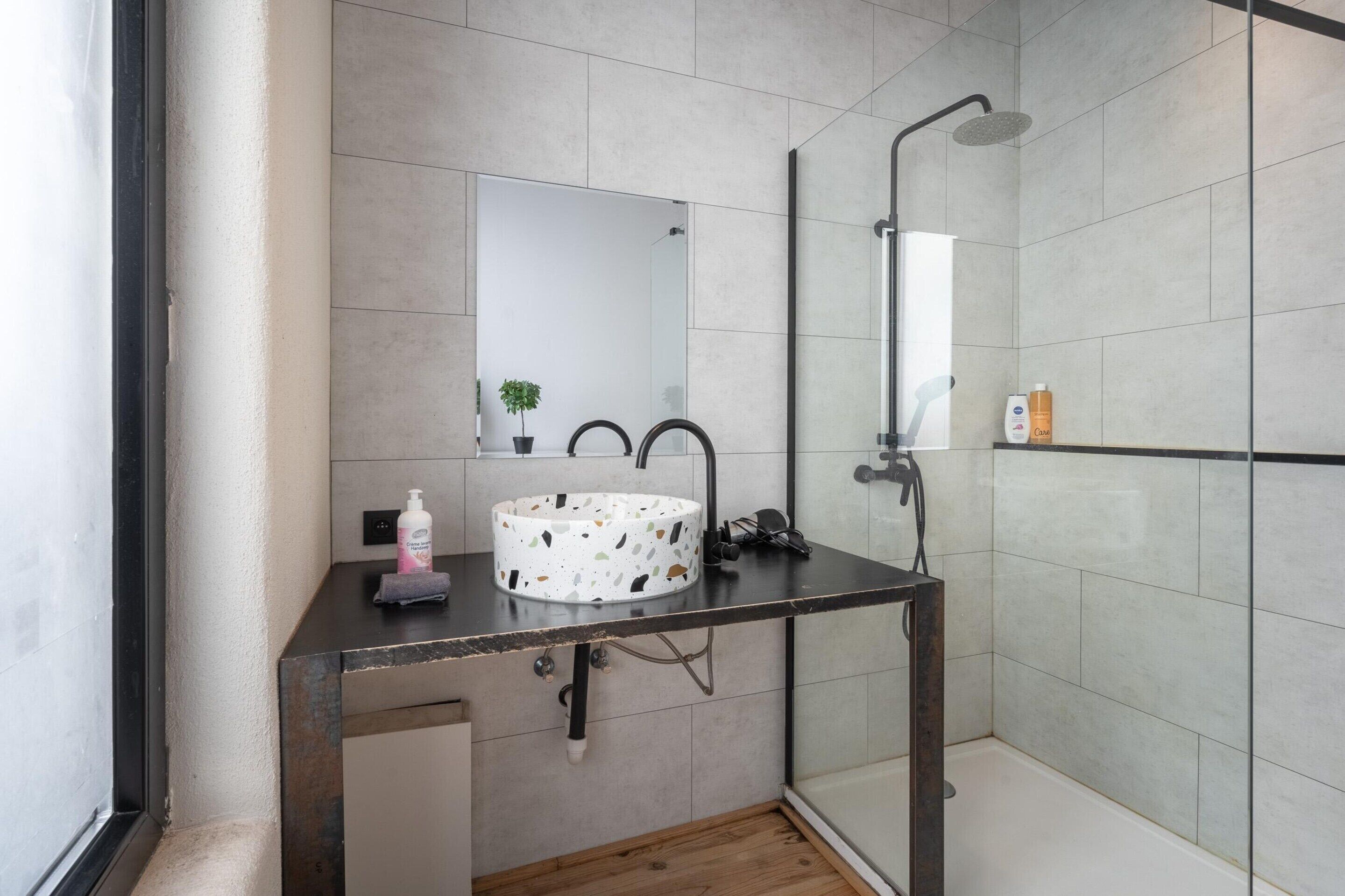 A modern bathroom with a round sink, a mirror, a shower, and a small potted plant.