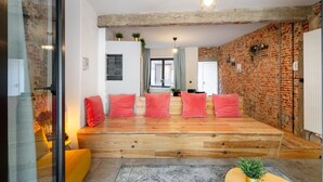 A modern living room with a wooden platform, red cushions, a glass partition, and a brick wall. - The Industrial Apartments 2bbknsinternet (Antwerp)