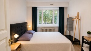 Ein ordentlich eingerichtetes Hotelzimmer mit Bett, Nachttisch, Lampe und Blick auf Bäume durch ein Fenster.
