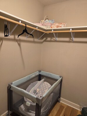 A corner of a room with a baby crib, a shelf with clothes, and a hanging rack with black hangers.