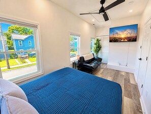A bedroom with a bed, a ceiling fan, a black leather sofa, a window with a view of a blue house, and a painting on the wall.