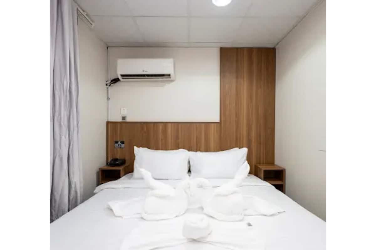 A hotel room with a bed, a wooden headboard, a nightstand, an air conditioning unit, and a curtain.