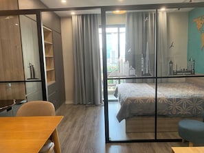 A hotel room with a bed, a desk, a chair, a wardrobe, and a view of city buildings. - Calmstay Luxury Apt Vinhomes D'Capitale  (Hanoi)