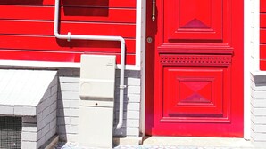 Eine rote Tür mit weißer Umrandung, ein Briefkasten und ein Verteilerkasten an einem Gebäude.