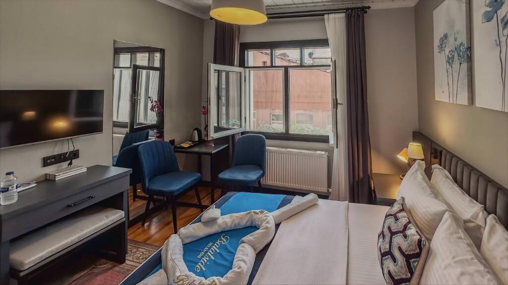 Egy szállodai szoba, amelyben található egy ágy, egy íróasztal, egy szék, egy televízió és egy ablak, amelyen függöny van.