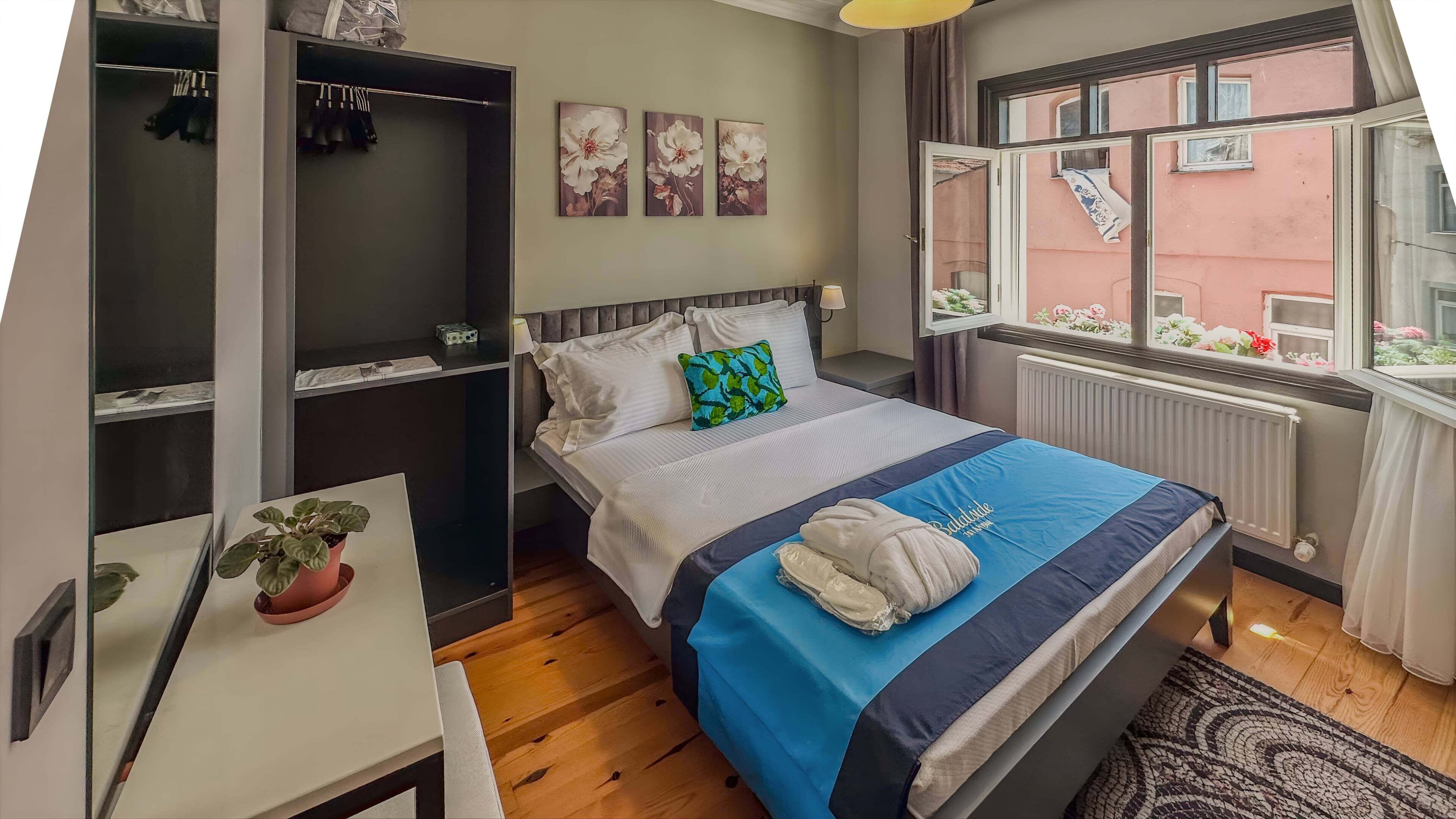 A bedroom with a bed, a desk, a potted plant, a wardrobe, and a window with a view of flowers.