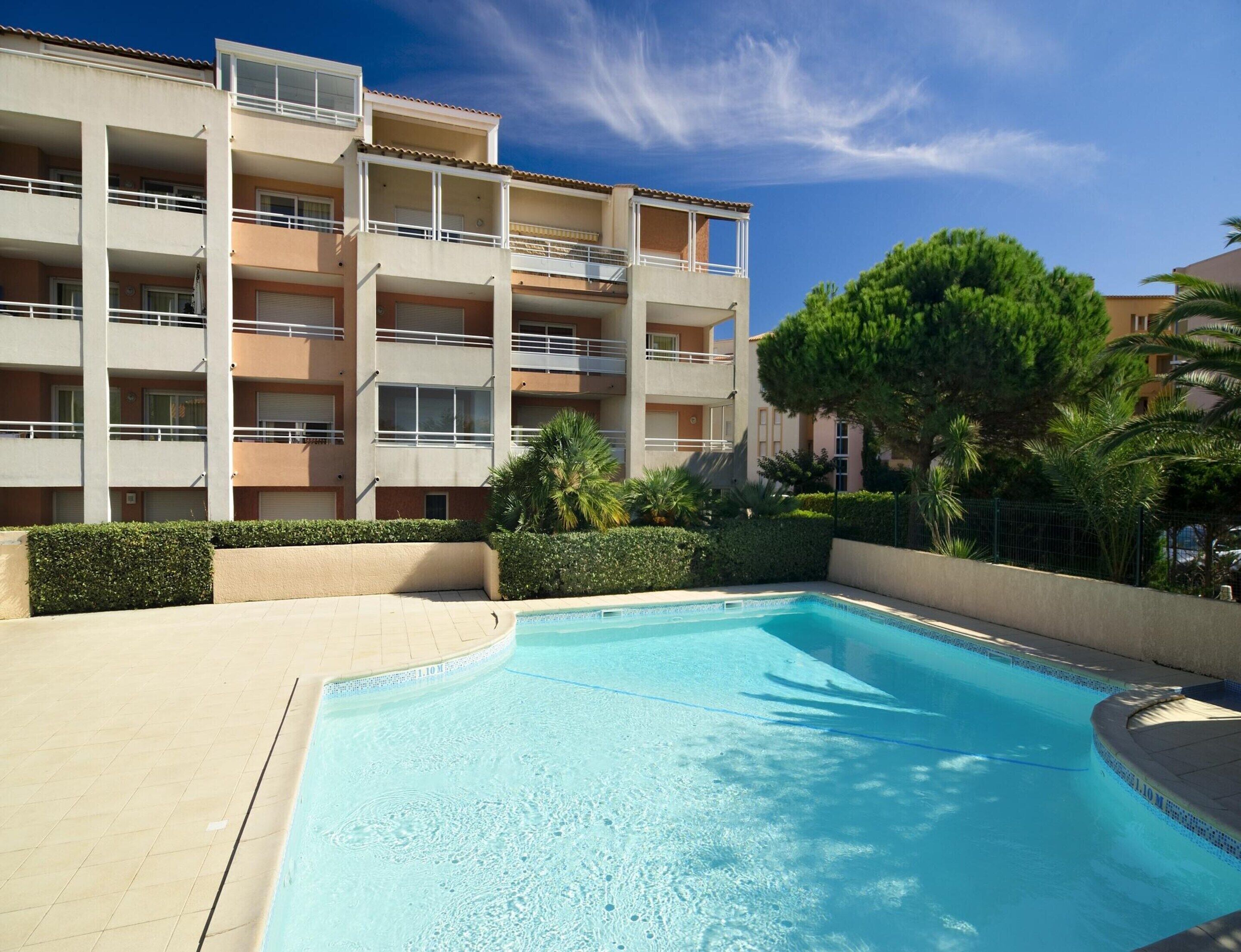 A swimming pool with a tiled edge, surrounded by a low hedge and a multi-story building with balconies.