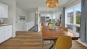 A spacious dining area with a wooden table and yellow chairs, a kitchen in the background, and a large window with curtains. - Marina-Front Retreat - Steps to Beach, Wine & Water (Algoma)