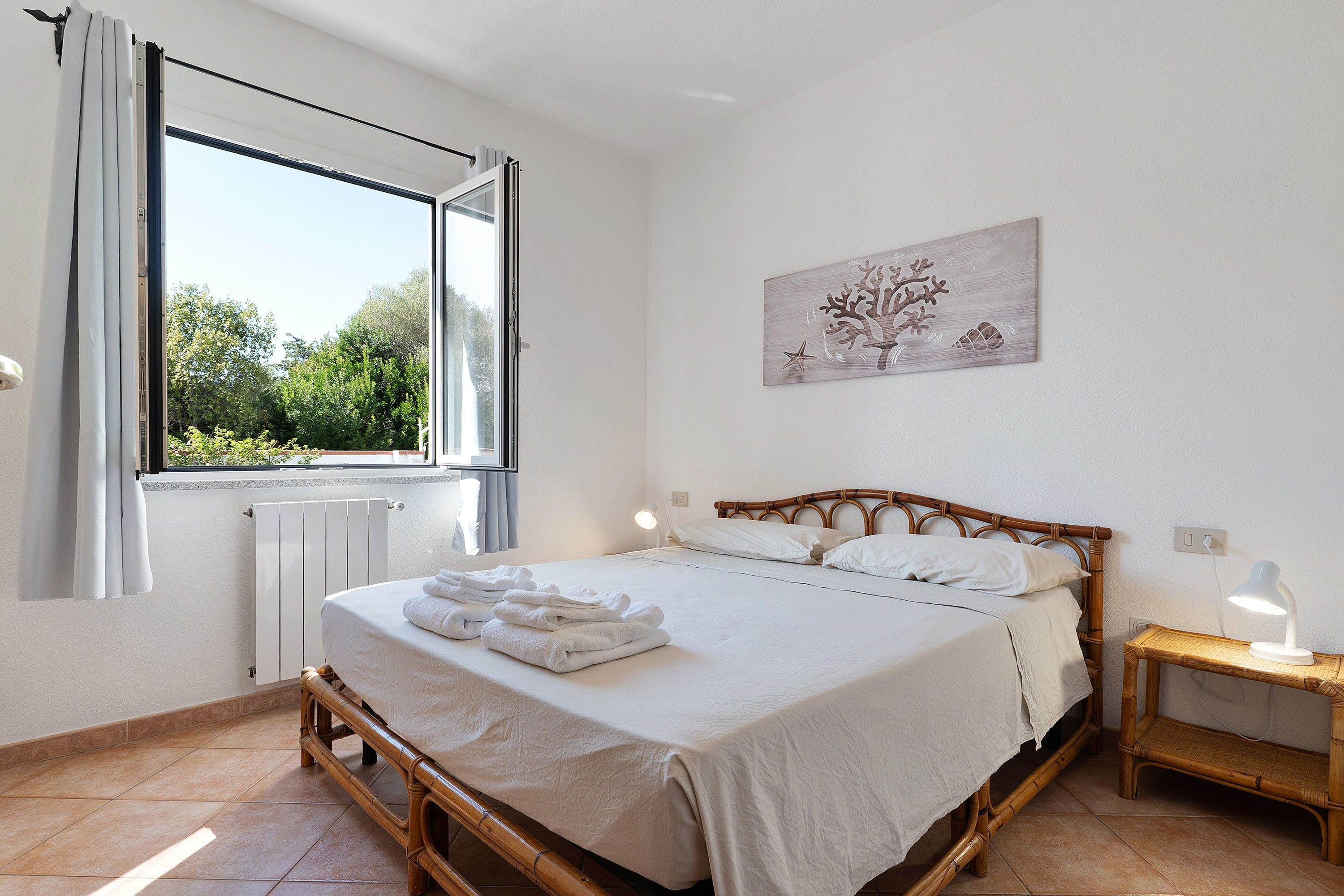 A bedroom with a bed, a bedside table with a lamp, a window with curtains, and a wall-mounted painting.