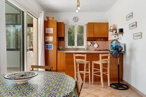 A compact kitchen with wooden cabinets, a dining table with chairs, a clock, and a fan. - Apartment 'Annina' with Private Terrace, Wi-Fi and Air Conditioning (Pittulongu)