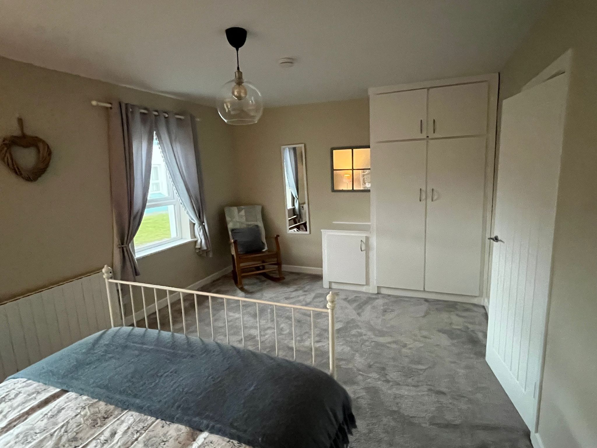 A bedroom with a bed, a rocking chair, a wardrobe, a small cabinet, and a window with curtains.