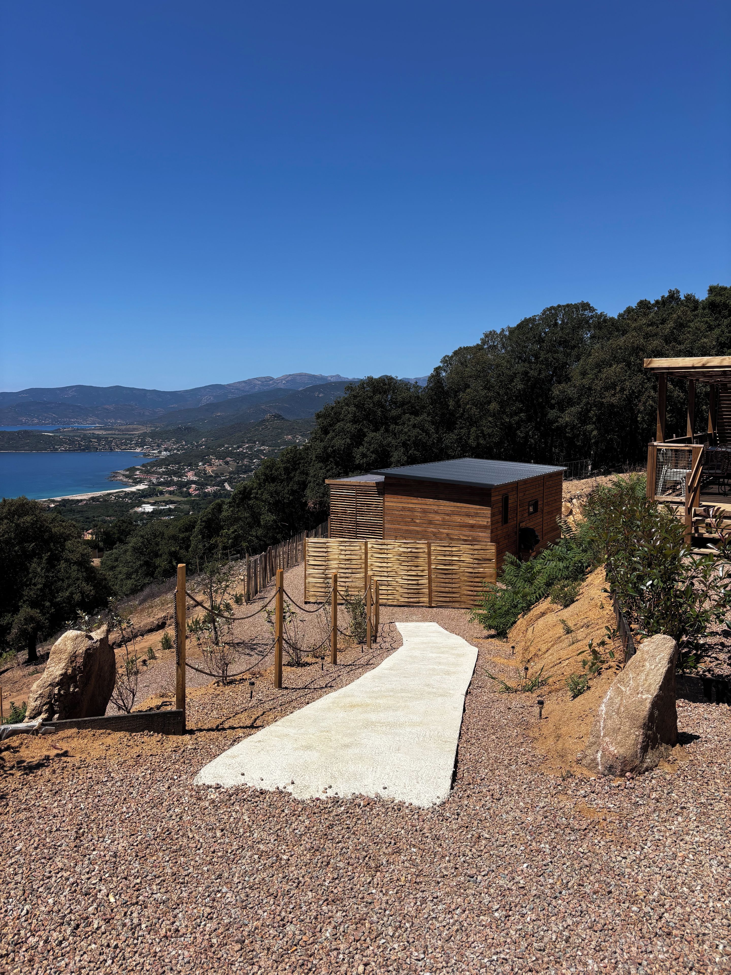 A modern building with a wooden exterior, a gravel pathway leading to it, and a view of a coastal landscape in the background.