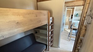 Un lit superposé avec un matelas foncé, une tête de lit en bois et un luminaire en matière tressée. Il y a une petite étagère sur le côté droit et une porte qui mène à un coin repas.