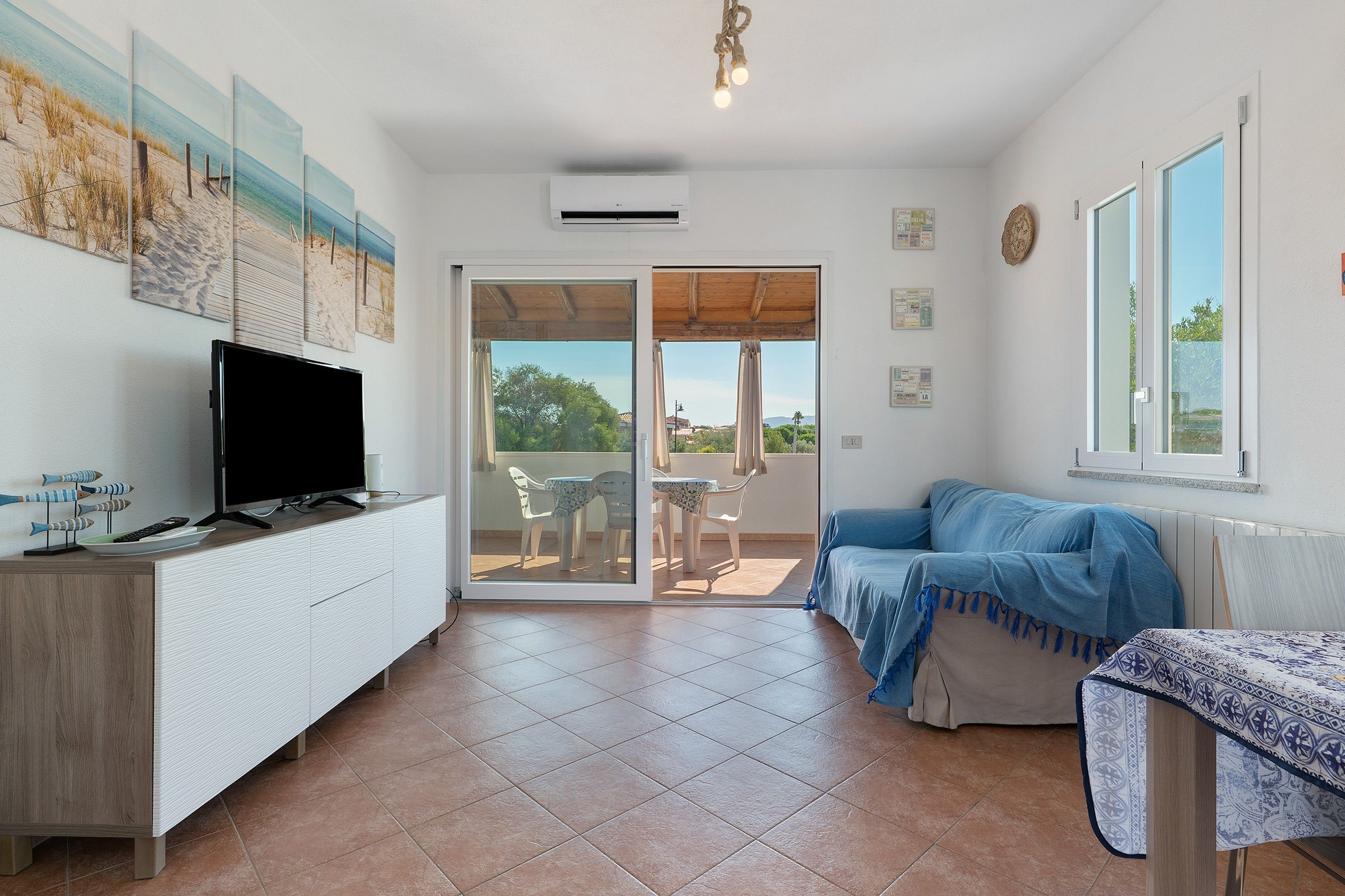 Une pièce avec un téléviseur posé sur un meuble blanc, un canapé bleu, une table recouverte d’une nappe à motifs et, à travers une porte vitrée, une vue sur un espace extérieur aménagé avec des chaises.