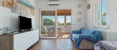 A room with a television on a white cabinet, a blue sofa, a table with a patterned tablecloth, and a view through a glass door to an outdoor area with chairs.