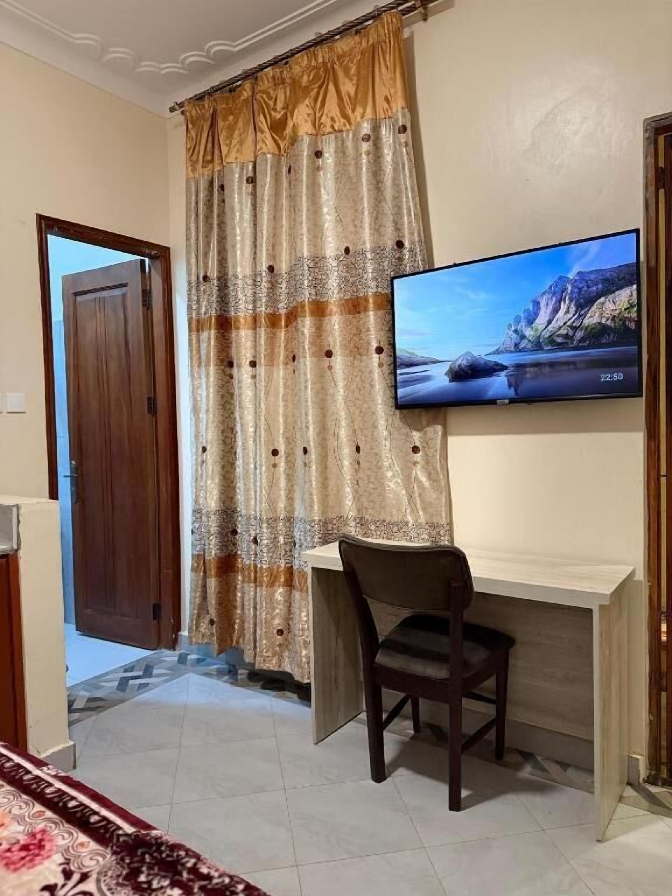 A room with a TV on the wall, a desk with a chair, a wooden door, and a patterned curtain.