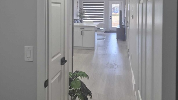 A hallway with a door on the left, a potted plant, and a window with blinds in the background. - Stylish Secondary Suite with Private Entry (Airdrie)