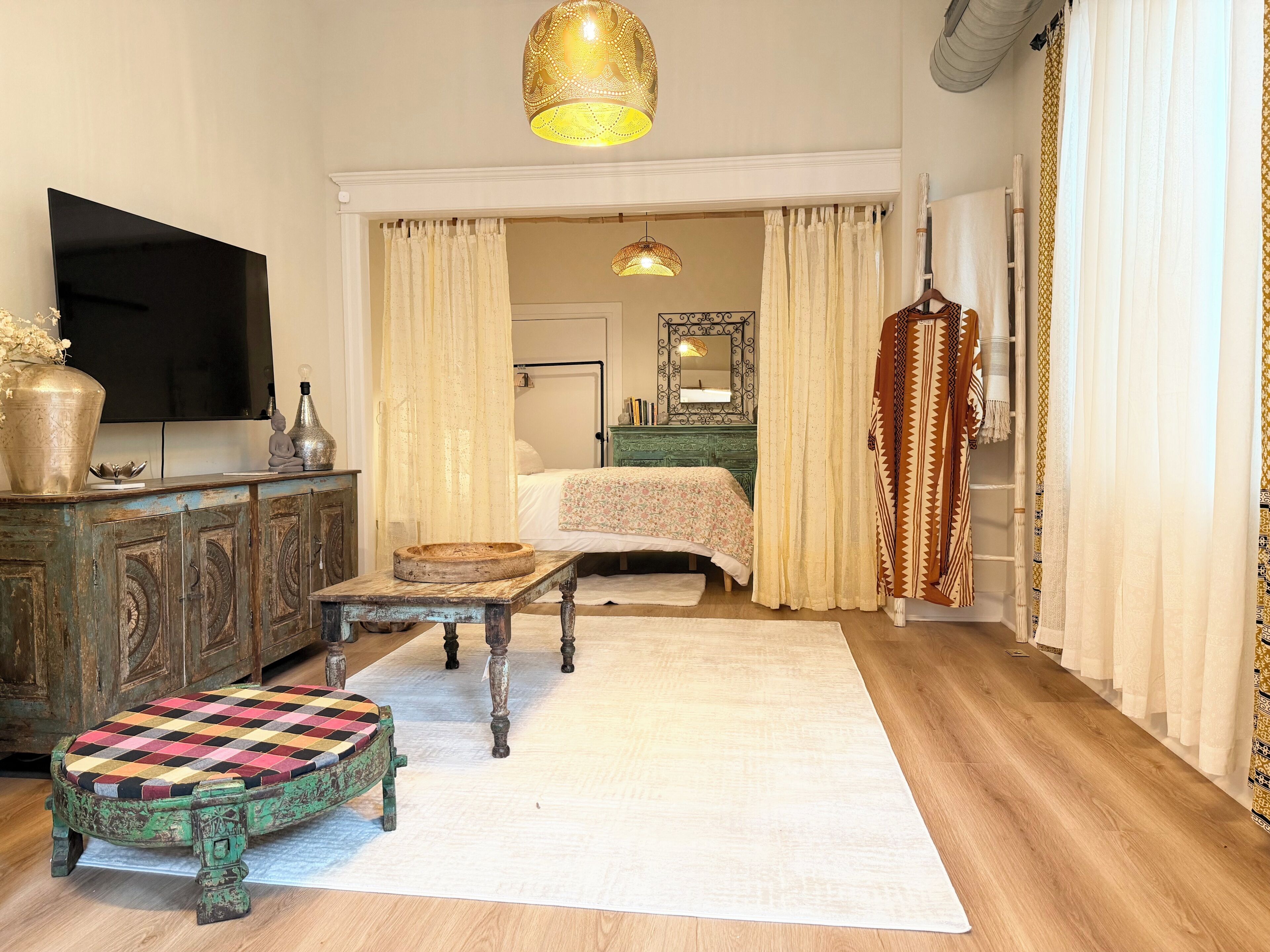 A room with a bed, a television, a wooden cabinet, a small table with a checkered cushion, and a hanging light fixture.