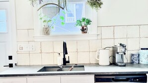 A kitchen with a stainless steel dishwasher, a sink, cabinets, a window with plants, and a hanging light fixture. - Light-Filled Boho Studio Alcove Retreat in Philly’s Best Neighborhood , new Deck (Philadelphia)