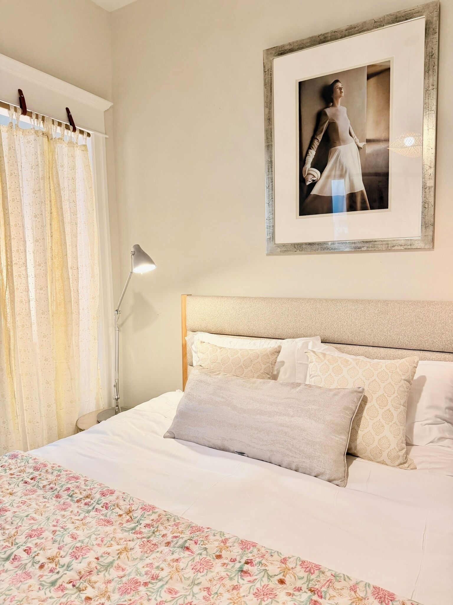 A bedroom with a bed, a headboard, a framed picture on the wall, and a window with curtains.