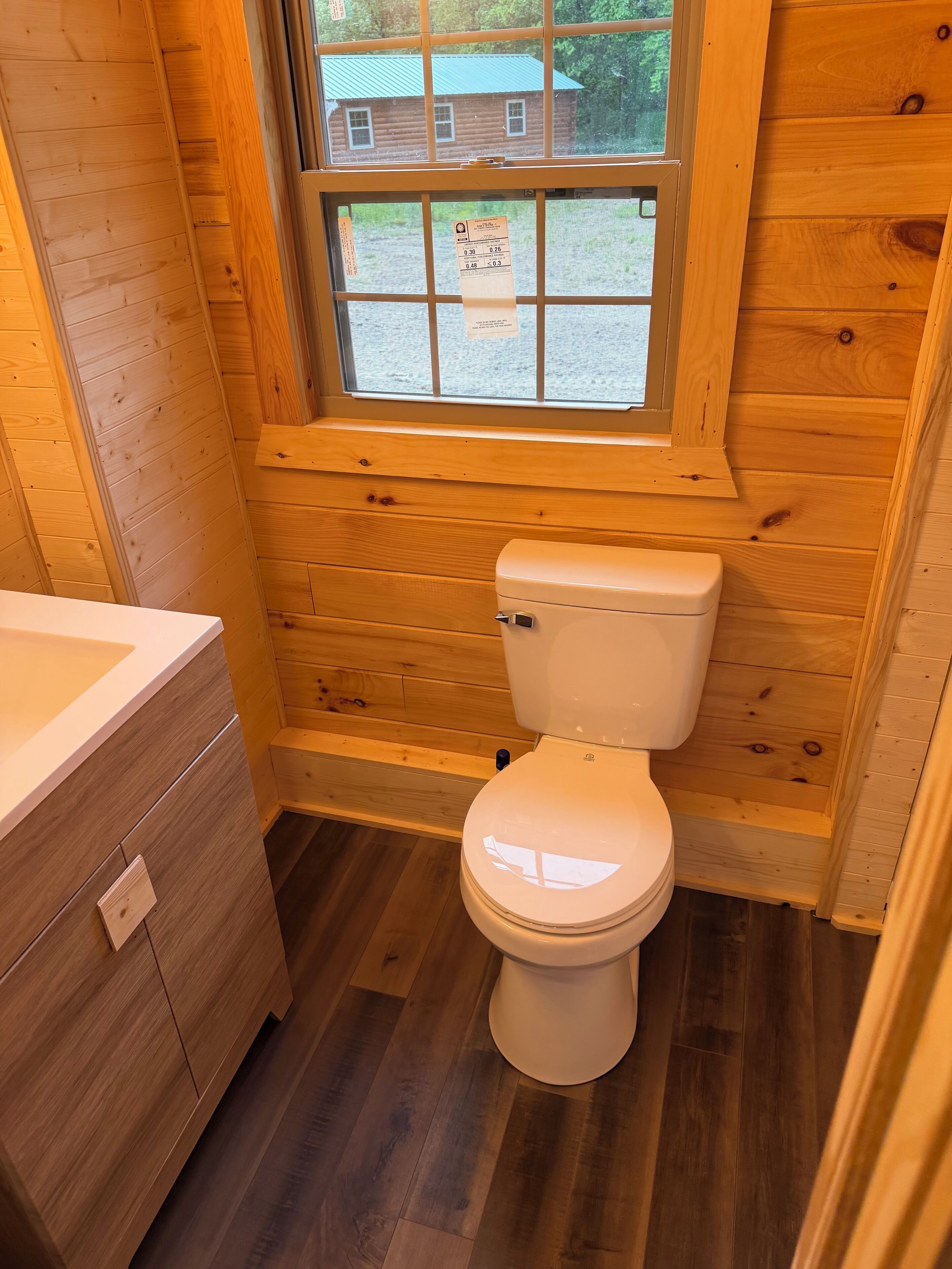 Een houten badkamer in een blokhut, met toilet, wastafel en raam met uitzicht op een huis.