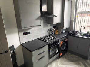 A modern kitchen with grey cabinets, a black countertop, a built-in oven, a washing machine, and a window with blinds.