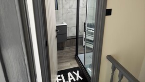 Ein modernes Badezimmer mit Duschkabine aus Glas, einem weißen Waschbecken und einer schwarzen Matte mit dem Aufdruck 'FLAX'.
