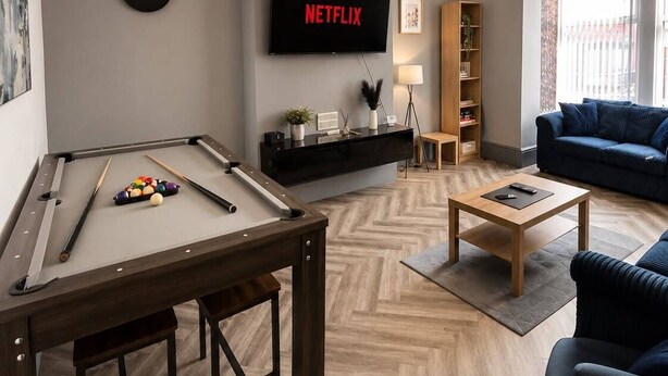 Ein modernes Wohnzimmer mit Billardtisch, einer Couch, einem Couchtisch, einem Fernseher, der 'NETFLIX' anzeigt, und einem Bücherregal.