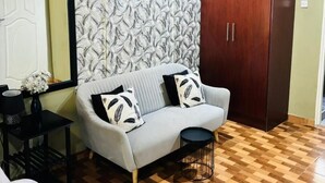 Een kamer met een houten plafond, een behang met patroon, een bank met gekleurde kussens, een donker houten garderobe en een vloerkleed met zebraprint.