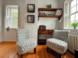 Une pièce avec un plancher en bois, deux fauteuils à motifs, un bureau avec une radio, des étagères et une fenêtre avec des volets.