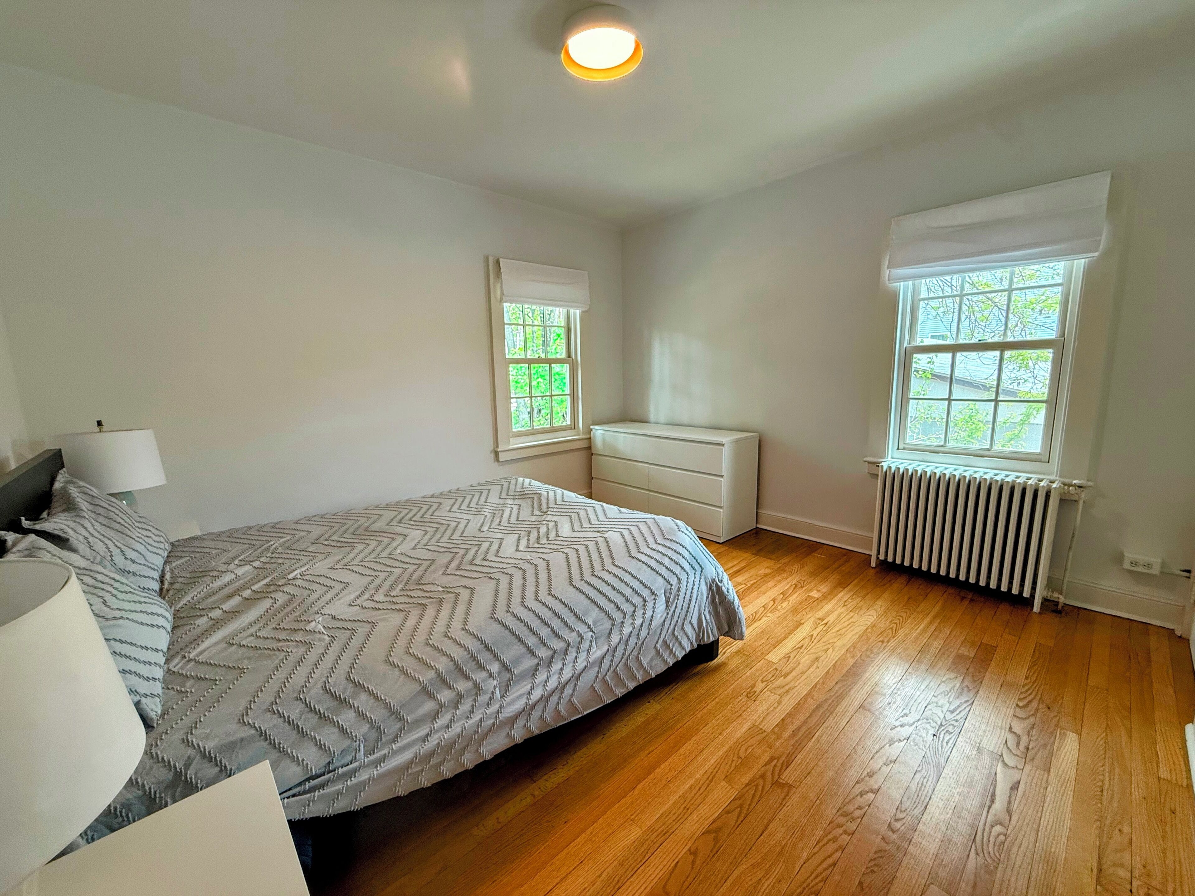 A bedroom with a bed, dresser, two windows, and a radiator.