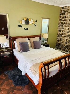 A bedroom with a wooden bed, a nightstand with a lamp, a wall clock, and a stone wall. - Studio in Golden Acres – Kwekwe - 2371 (Kwekwe)