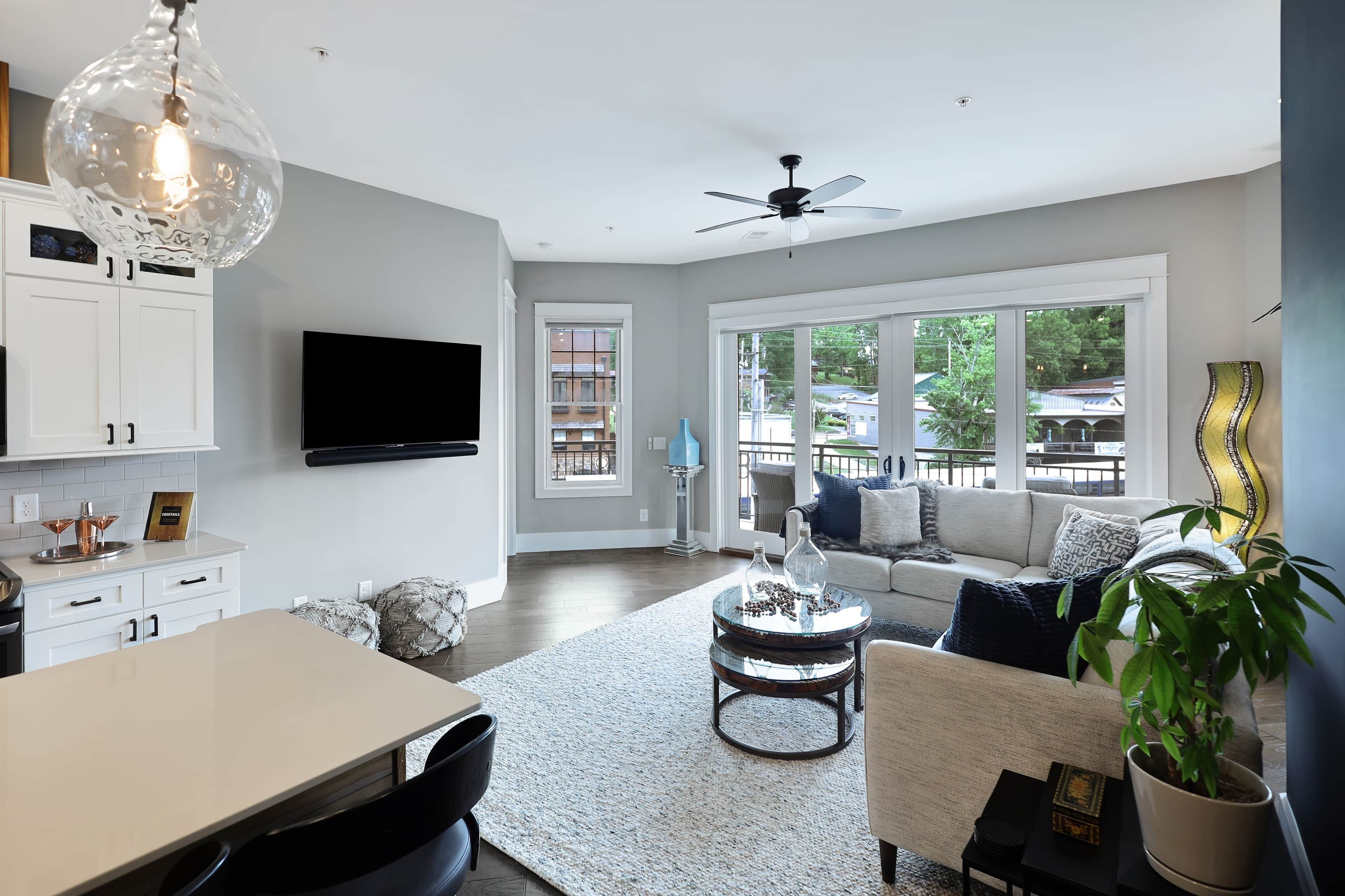 A modern living room with a dining area, a ceiling fan, a flat-screen TV, and large windows.
